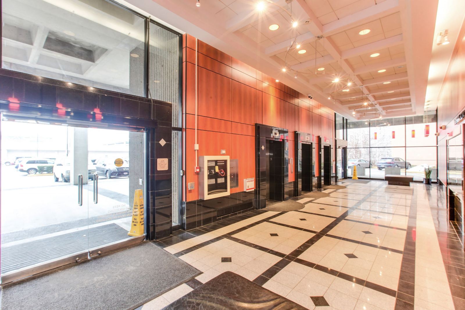Before: the original lobby seen from the glazed entrance doors, with orange-brown veneer panelling running the full wall past the elevator entrances, surface-mounted fire and control panels, beige patterned floor tile and track lighting on a coffered ceiling.