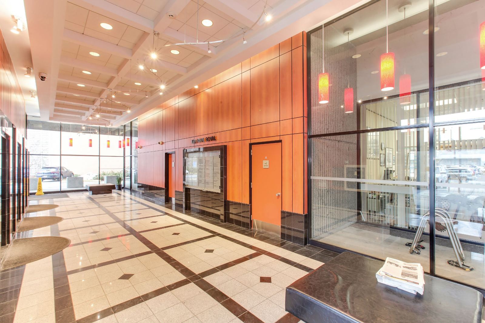 Before: the original 100 Park Royal lobby looking down the length of the space, with dated orange-brown wood veneer wall panelling, a printed tenant directory board, beige and brown patterned floor tile, a coffered white ceiling and red and orange glass pendant lights.