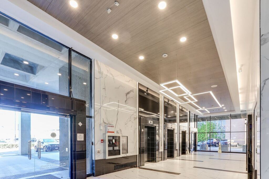 Upward view of the new millwork ceiling feature in timber-effect panels, with recessed downlights and two suspended interlocking square LED fittings running along the lobby toward the elevator bank.