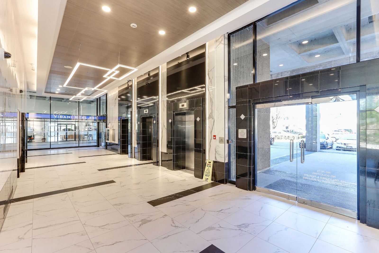Wide view of the finished lobby with three black-clad elevator entrances on the right, stainless doors and diamond inlays, timber-effect ceiling with square LED pendants, and the glazed street doors beyond.