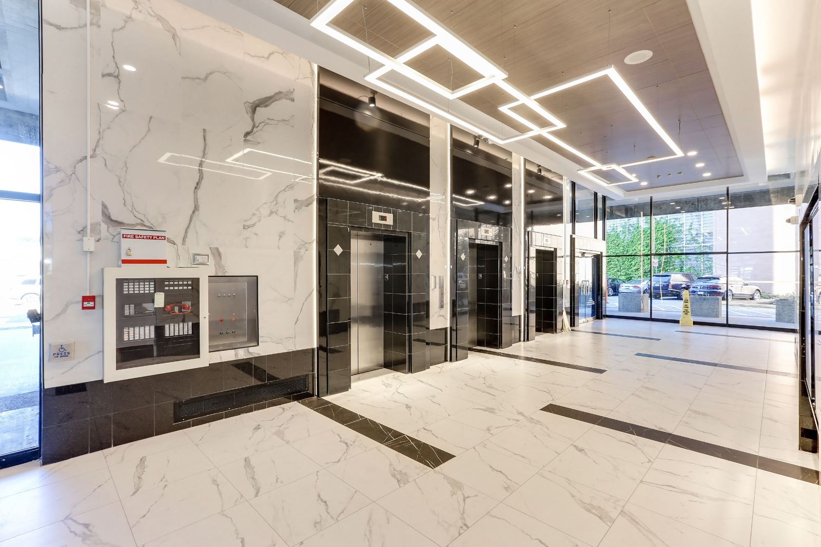 Side angle along the elevator bank showing large-format marble-look porcelain wall panels, the recessed fire safety and control panels set flush into the wall, black polished tile base with a floor grille, and square LED pendants overhead.