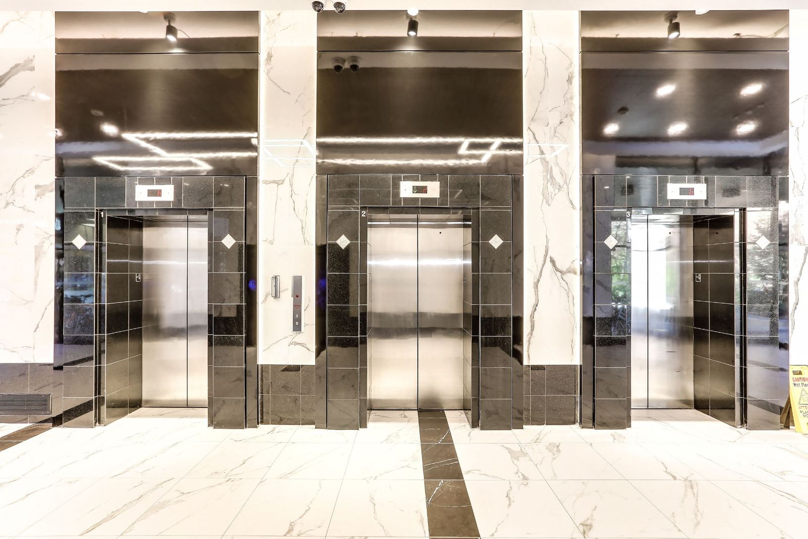 Straight-on view of three elevator entrances numbered one to three, each framed in polished black tile with white diamond inlays and digital floor indicators, separated by marble-look porcelain panels above brushed stainless doors.
