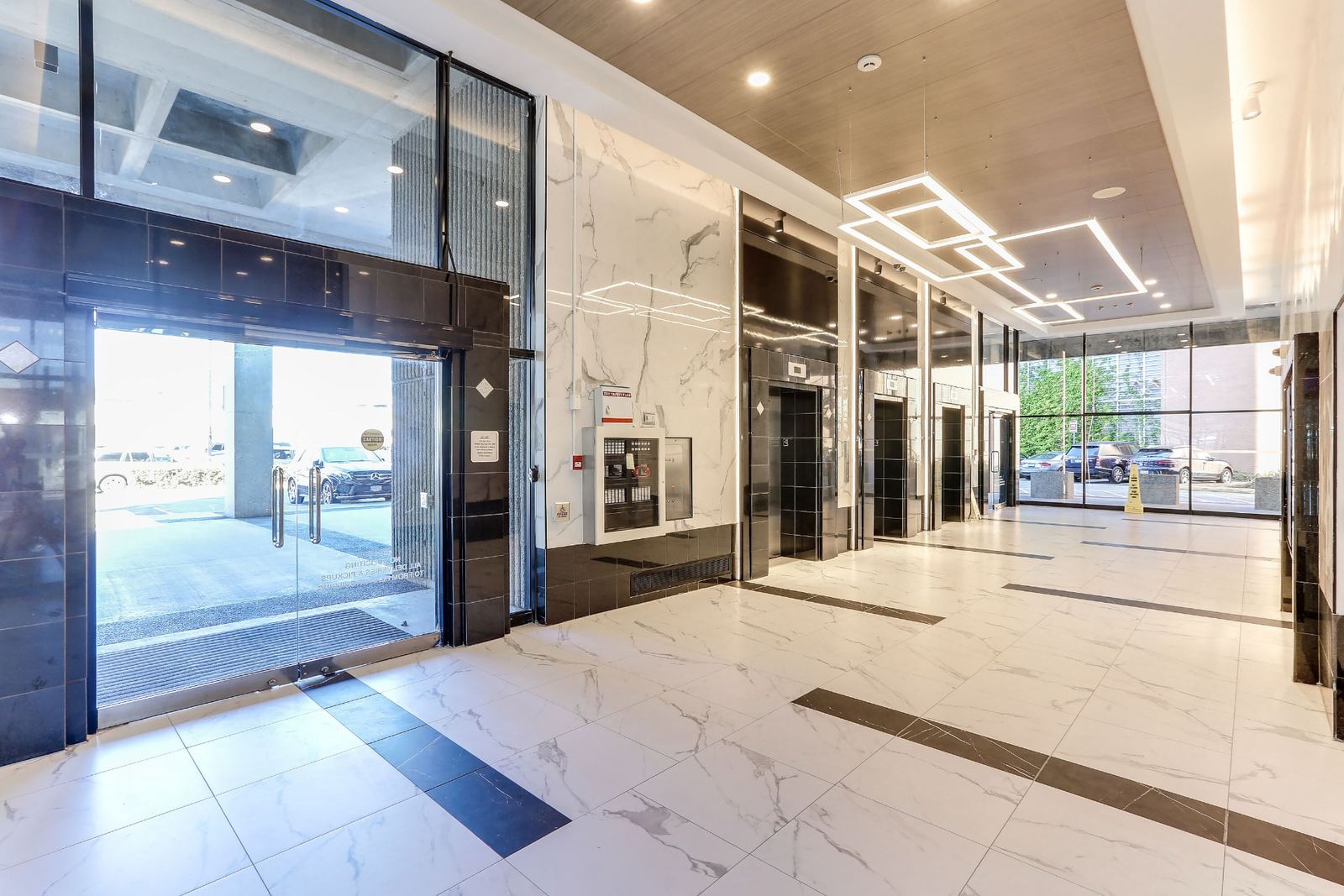 View across the finished lobby to the glass entrance doors, with a black polished tile entrance surround, marble-look porcelain wall panels carrying a recessed lit reveal, black tile base and the elevator bank receding to the right.