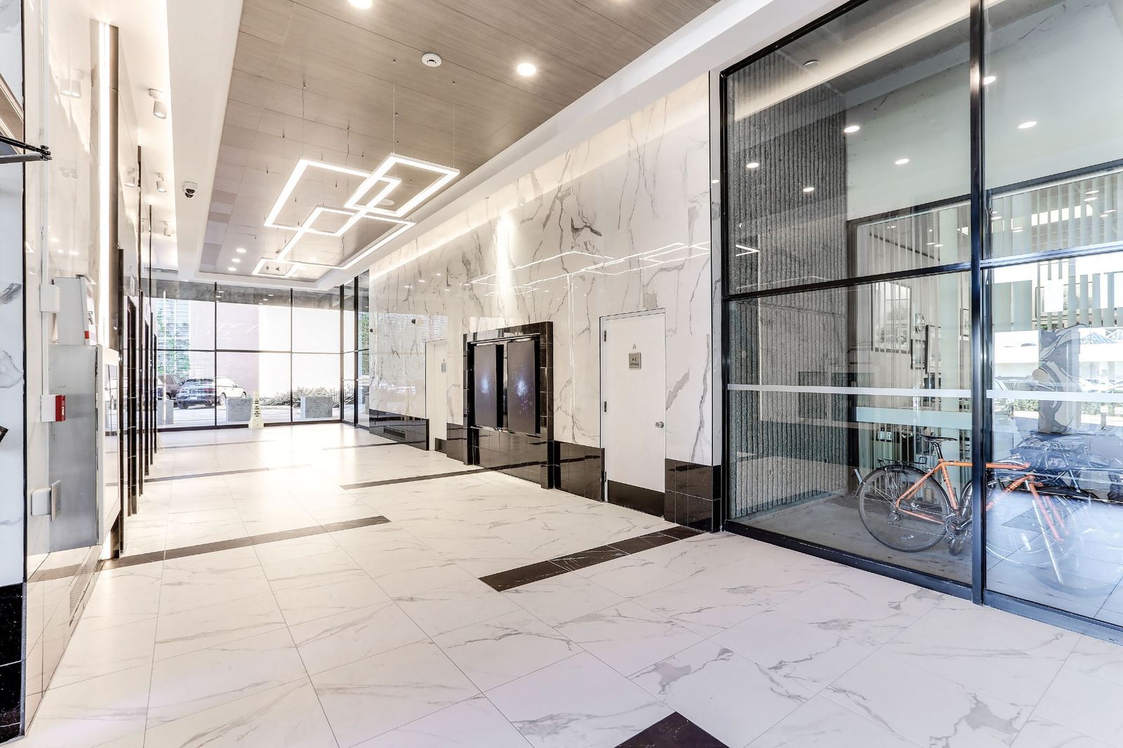 Renovated 100 Park Royal lobby looking down the full length of the space, with marble-look porcelain wall tile, a white marble-pattern tiled floor banded in black, overlapping square LED ceiling pendants and a glazed bicycle storage room to the right.